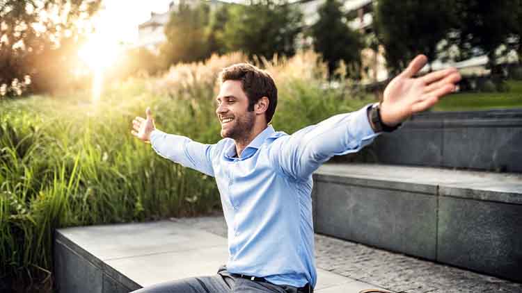 A happy young businessman with arms stretched outdoors at sunset.