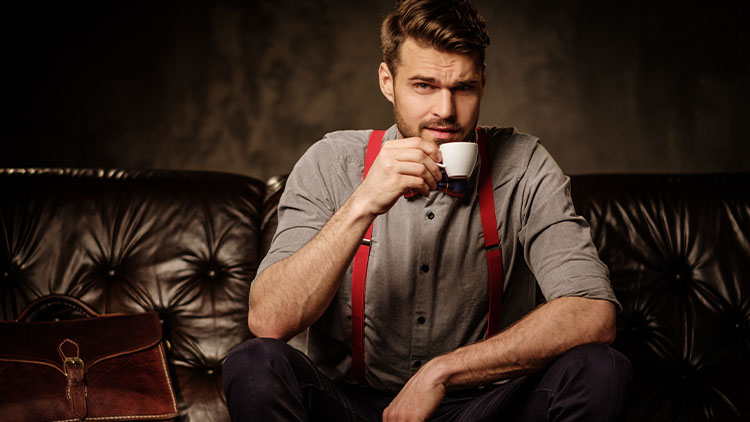 Young handsome old-fashioned bearded man with cup of coffee sitting on comfortable leather sofa on dark background.
